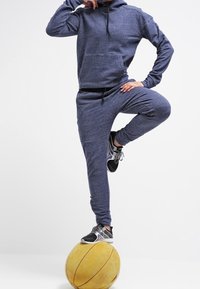 Sweat à capuche bleu marine et joggers assortis en tissu texturé, avec une poche avant et un cordon de serrage. L'athlète se tient debout sur un ballon de basket jaune.