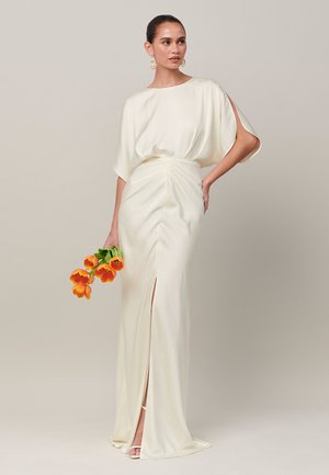 Frau in langem weißen Kleid mit Schlitz, die orange Tulpen hält, vor schlichtem beigem Hintergrund stehend.
