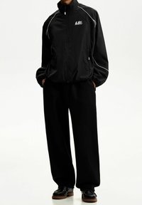 Zwarte windbreakerjas met witte accenten, lichtgewicht materiaal, hoge kraag en zijzakken. Gecombineerd met loszittende zwarte broek.