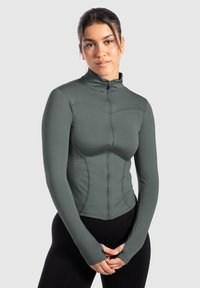 Chaqueta ajustada de manga larga en verde oliva con cremallera frontal, cuello alto y agujeros para los pulgares. Presenta detalles cosidos.