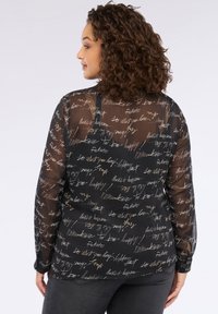 Donna con capelli ricci che indossa una blusa nera trasparente a maniche lunghe con testo scritto a mano bianco e beige, abbinata a pantaloni scuri, vista da dietro.