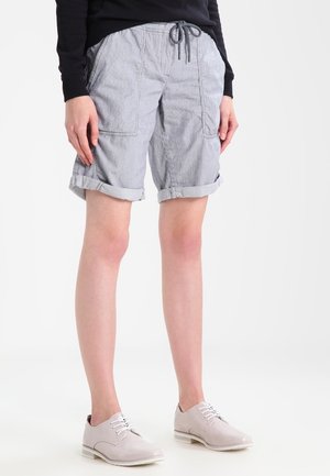 Person trägt graue, hochgekrempelte Shorts bis zum Knie mit großen Vordertaschen, ein schwarzes Langarmshirt und hellbeige Schnürschuhe.