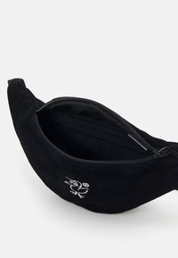 Svart kordfløyelsfanny pack med rund fasong, glidelås-lukking, og et brodert fugledesign foran. Innvendig har den en separat lomme.