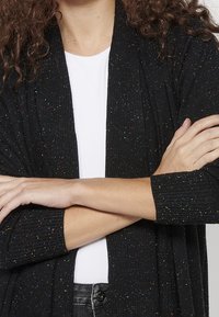 Svart öppenstickad cardigan med flerfärgade prickar, ribbad textur, trekvartsärmar, bärs över en vit topp, med armarna korsade.