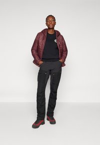 Giacca trapuntata rosso scuro con cappuccio, felpa nera con logo, pantaloni cargo neri con tasche laterali e scarponi da trekking rossi e neri.