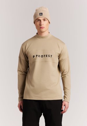 Young man wearing beige beanie and matching long sleeve shirt with "Protest" text, paired with black pants, standing against plain background.