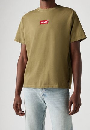 Homme portant un T-shirt Levi's vert olive avec un petit écusson logo rouge sur la poitrine et un jean en denim bleu clair.