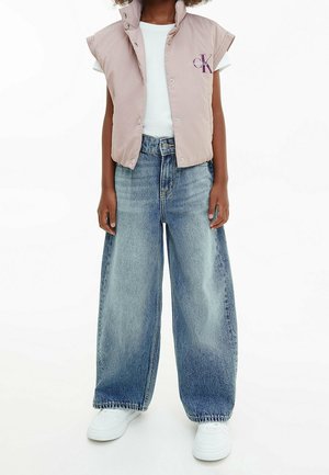 Person iført lyserød let uden ærmer dunvest over hvid t-shirt, vidde falmede blå jeans og hvide sneakers, stående på hvid baggrund.