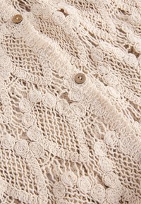 Pitskootud beež rõivas, millel on tekstuuriga lillemustrid ja mis on kinnitamiseks lõpetatud puidust nööpidega. Kerge ja õhuline disain.