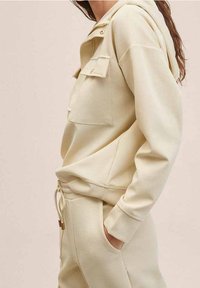 Helles beige Kapuzensweatshirt mit zwei Brusttaschen und gerippten Bündchen. Hergestellt aus weichem Material mit lockerer Passform und verstellbarem Kordelzug.