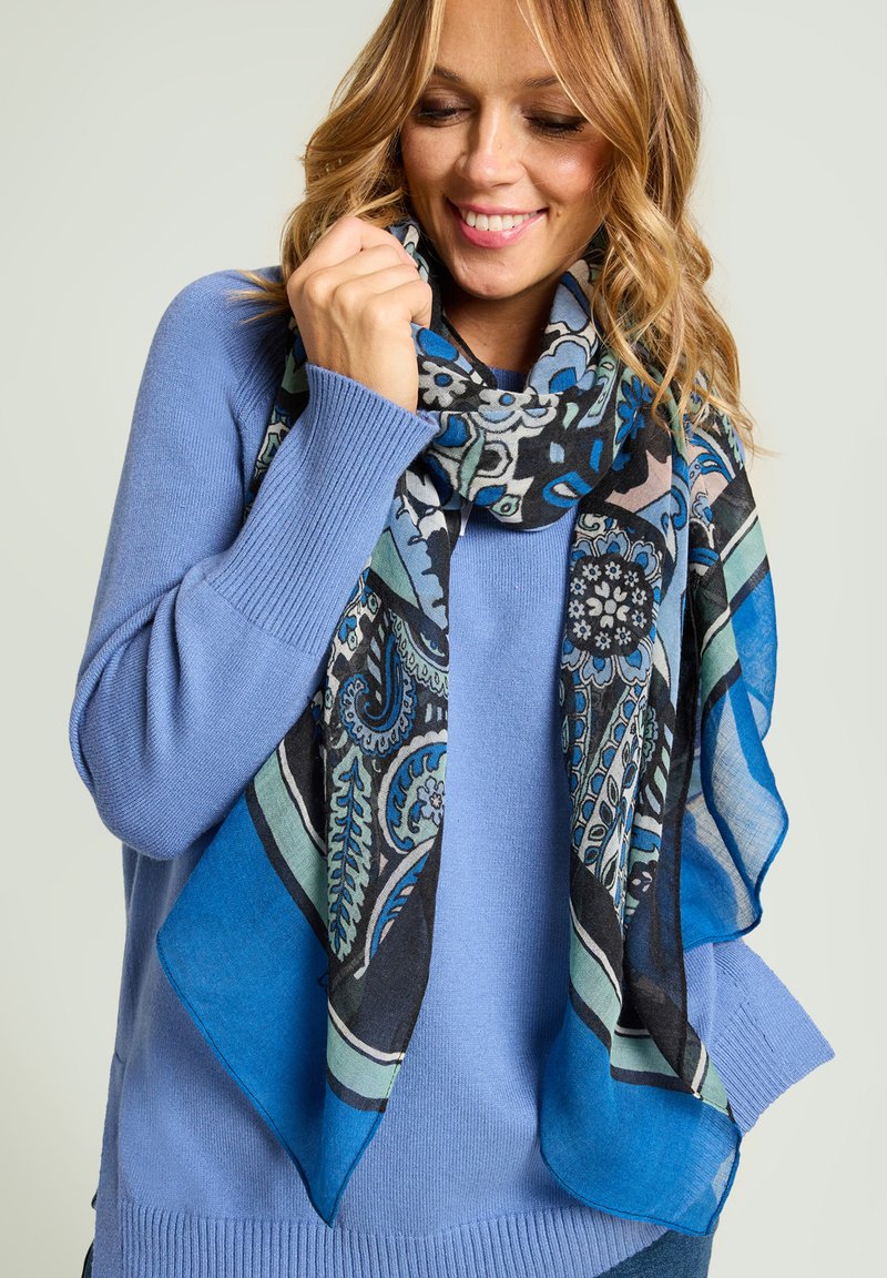 Écharpe noire et bleue à motifs, drapée autour d'un pull bleu, présentant des designs complexes et un tissu léger et transparent.