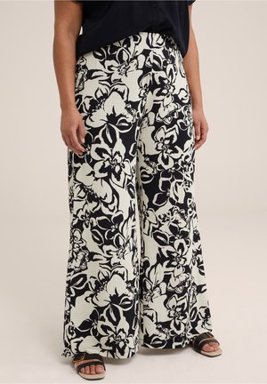 Personne portant un pantalon large avec un grand imprimé floral noir et blanc, un haut noir et des sandales noires avec des semelles marron clair.