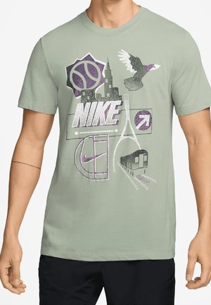 Mężczyzna noszący jasnografitową koszulkę z logo Nike, z grafikami panoramy miasta, koszykówki, ptaka, pociągu i strzałki kierunkowej na przodzie.