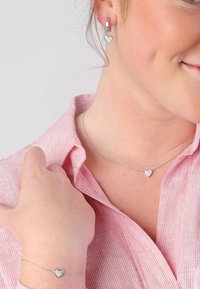 Zilveren sieradenset met een hartvormige hangerketting, bijpassende armband en oorbellen met gestructureerde details. Gedragen door een model in een gestreept shirt.