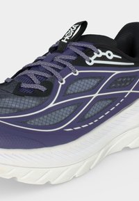 Κοντινή εικόνα ενός μωβ και μαύρου αθλητικού παπουτσιού με λευκή σόλα, πάνελ με δίχτυ, κορδόνια με σχέδια και λογότυπο HOKA στην γλώσσα του παπουτσιού.