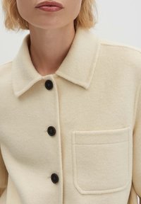 Giacca in lana beige chiaro con collo a risvolto, bottoni neri e taschino sul petto. La trama del tessuto è morbida e leggermente pelosa.