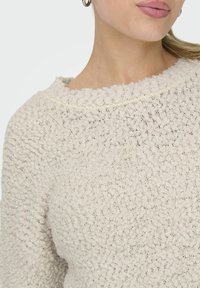 Maglione di un beige chiaro con texture e collo tondo. Il tessuto presenta un motivo a bolle e un delicato lucido. Finitura liscia sui bordi.