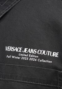 Černá látka s bílým výšivkovým textem: "VERSACE JEANS COUTURE Limited Edition Fall Winter 2025 2026 Collection." Hladký povrch, žádné vzory.