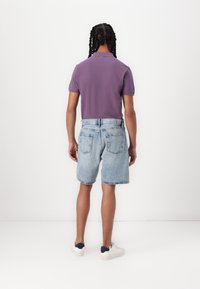 Camisa polo morada de manga corta combinada con pantalones cortos de mezclilla azul claro, con un dobladillo deshilachado y dos bolsillos traseros. Zapatillas blancas.