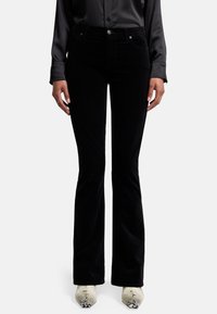 Jeans en velours côtelé noir à jambes évasées, avec un design à cinq poches, fermeture à bouton à l'avant et tissu texturé, associés à des bottes pointues de couleur claire.