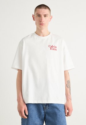 Camiseta blanca de algodón con cuello redondo, mangas cortas, y un logo rojo de "Calvin Klein" impreso en el lado izquierdo del pecho. Llevada con jeans de mezclilla azul.