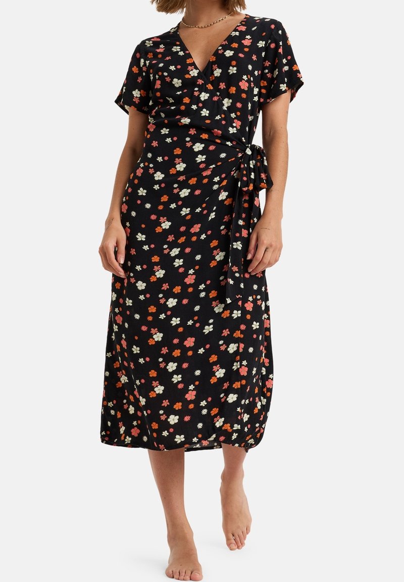 Femme en robe portefeuille noire à manches courtes avec un petit imprimé floral multicolore, debout pieds nus sur fond blanc.