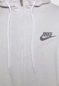 Světle šedá mikina na zip vyrobená z měkké tkaniny, s krátkým zipem, bílými šnůrkami a třpytivým logem Nike na hrudi.