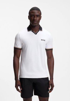 Mężczyzna ubrany w biały polo z czarnym kołnierzem i logo „BOSS”, w zestawieniu z czarnymi szortami na jednolitym białym tle.