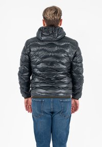Giacca a vento in materiale nero lucido con cappuccio, caratterizzata da onde trapuntate e orlo aderente. Indossata sopra jeans blu.