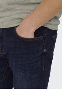 Persona con la mano parcialmente en el bolsillo de unos pantalones vaqueros de mezclilla azul oscuro, vistiendo una camisa texturizada de color verde claro.