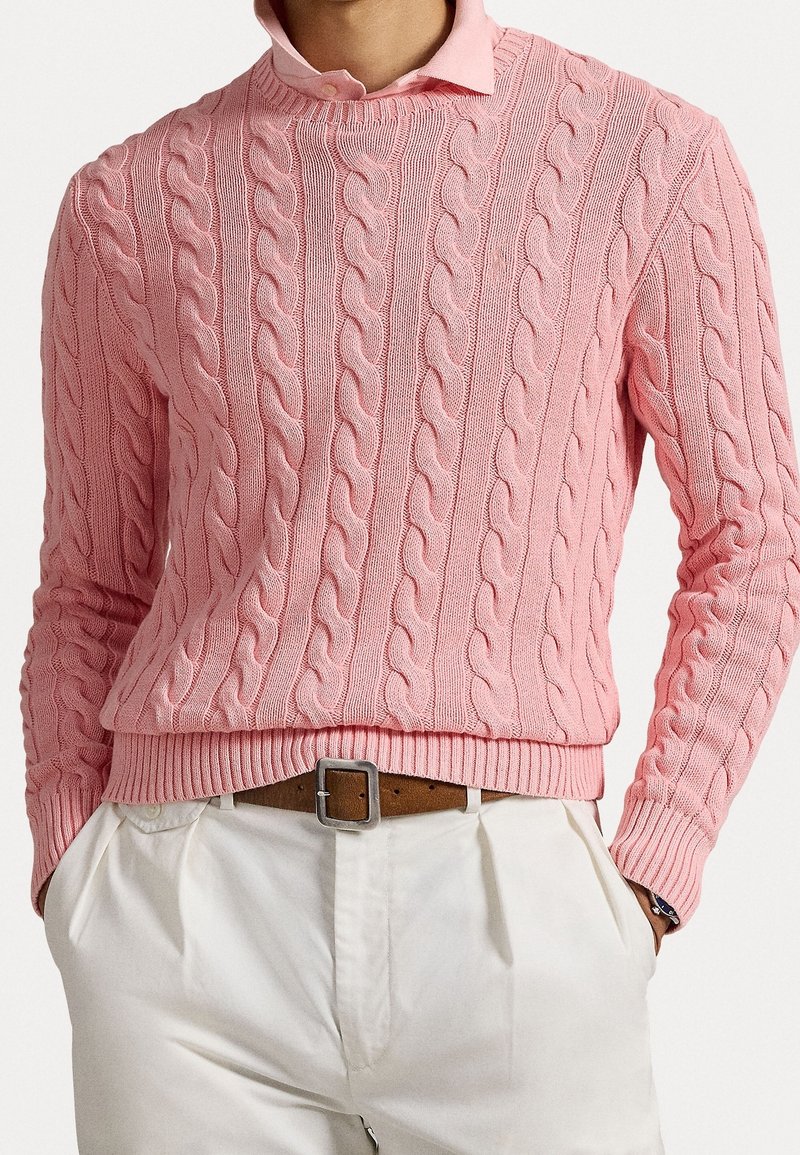 Homme portant un pull torsadé rose par-dessus une chemise rose clair à col, un pantalon blanc plissé et une ceinture en cuir marron avec une boucle argentée.