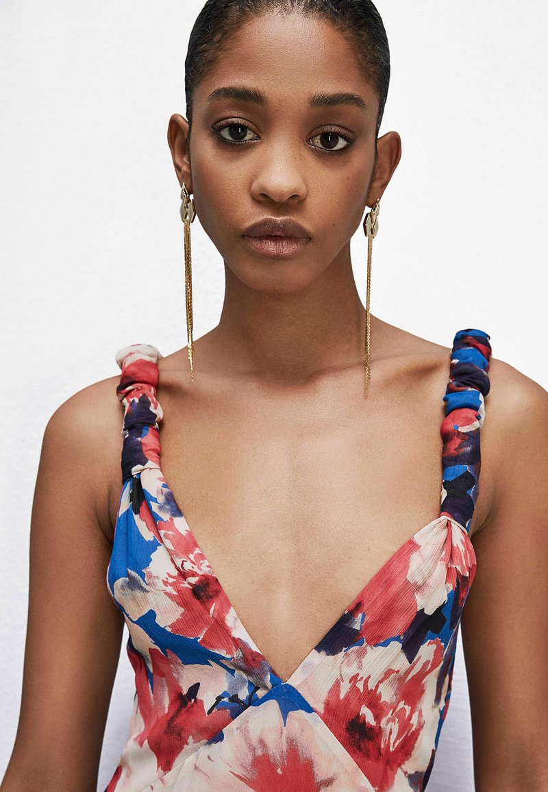 Abito floreale con design a V, caratterizzato da motivi blu, rossi e bianchi. Le spalline sono intrecciate con una finitura texture. Orecchini a cerchio dorati.