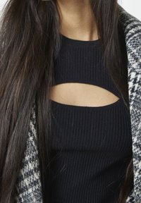 Musta ribbineuloksinen hihaton paita, jossa on leikkausdetaili, yhdistetty harmaaseen kuviolliseen neulecardiganiin. Pitkät tummat hiukset peittävät osittain asun.