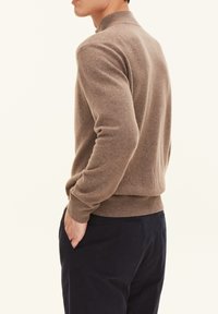Maglione di colore marrone a maglia con colletto alto, polsini e orlo a costine. La texture sembra morbida e leggermente pelosa, indossato con pantaloni scuri.