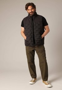 Gilet rembourré noir avec un col montant, doté d'un design matelassé, porté par-dessus une chemise noire. Associé à un pantalon vert olive et des baskets beiges.
