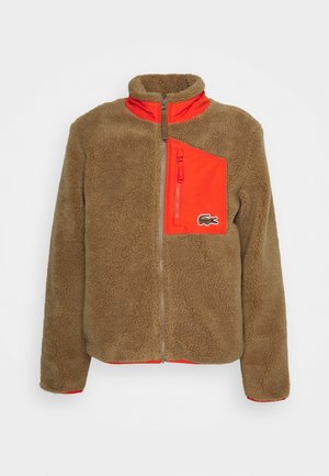 Veste polaire marron avec une poche poitrine et des accents de col orange vif, fermeture éclair frontale et petit logo crocodile sur la poche.