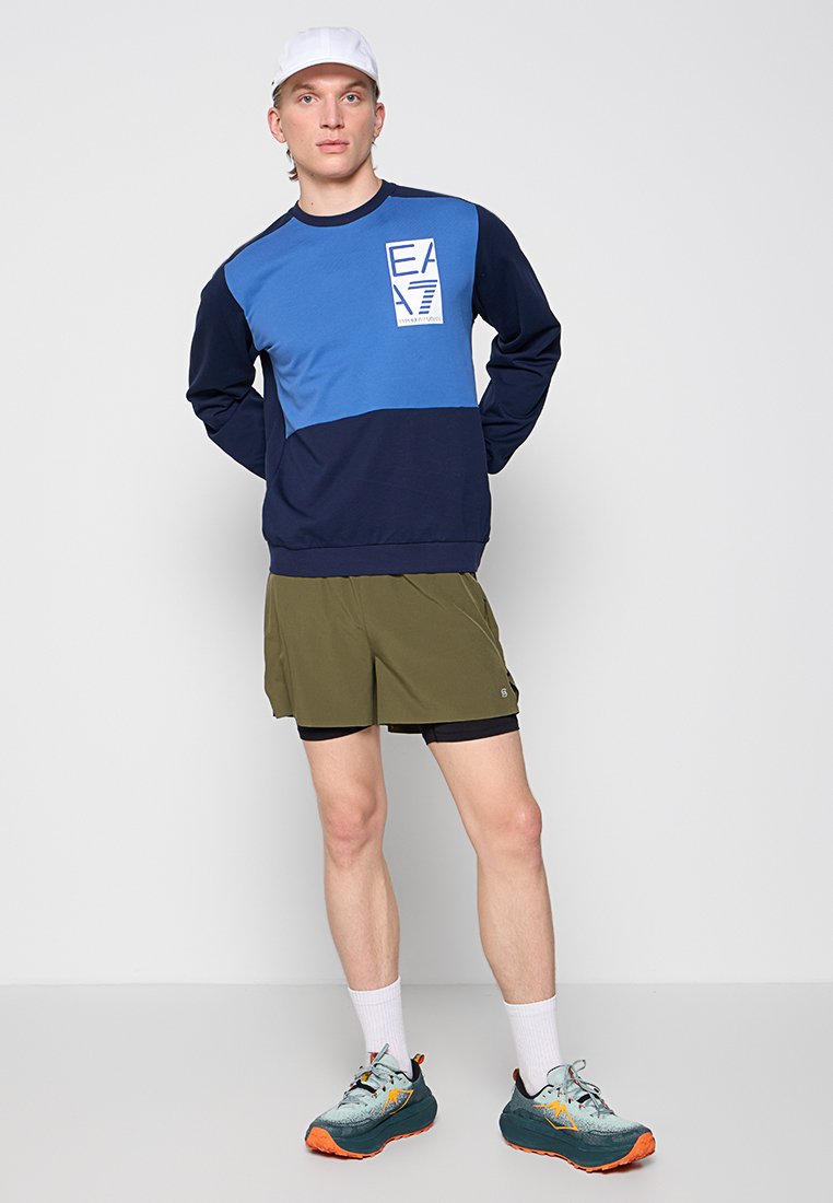 Sudadera azul y marino con logo, combinada con pantalones cortos verde oliva. El modelo lleva calcetines blancos y zapatillas deportivas turquesas con acentos naranjas.