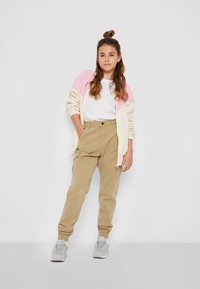 Lättvikts zip-up hoodie i krämfärg med rosa accenter, kombinerad med beige cargobyxor och grå sportskor. Modellen står med händerna på höfterna.