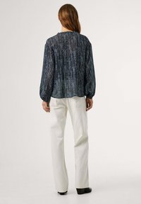 translucente, donkerblauwe blouse met verticale strepen, geplooide halslijn en pofmouwtjes, gecombineerd met witte broek en zwarte hakken.