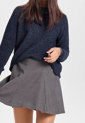 Kobieta w ciemnoniebieskim dzianinowym swetrze i czarno-białej spódnicy w kratę, trzymająca brzeg swetra jedną ręką.