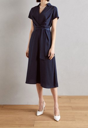 Kobieta w granatowej sukience kopertowej z krótkimi rękawami, z paskiem zawiązanym w talii, białe szpilki, perłowy naszyjnik i pierścionek.