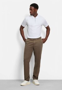 Witte poloshirt met een halve rits aan de hals, gecombineerd met khaki chino's. Model draagt witte sneakers; staande houding, handen in de zakken.