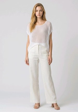 Joven con cabello largo y rubio, vistiendo una camiseta blanca de punto de manga corta, pantalones blancos de pierna ancha y talle alto, y sandalias beige de tacón.