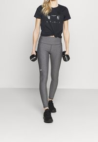 Mujer con camiseta negra de Under Armour y leggings grises sosteniendo pesas de 3 kg, de pie sobre un fondo claro y uniforme.