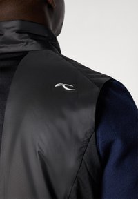 Čierny, ľahký vest s hladkou textúrou a vysokým golierom. Obsahuje biele logo na pleci a kontrastnú modrú látku pod ním.