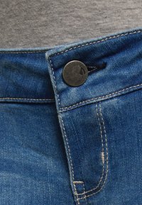 Os jeans de denim azul apresentam um botão de metal, costura em contraste e uma textura suave. A cintura é visível acima dos jeans, mostrando um tecido cinza.