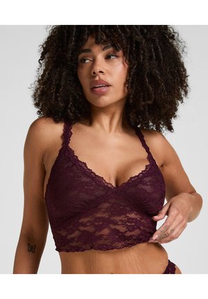 Woman with curly hair wearing a dark purple lace bralette, slightly pulling the fabric near her waist, showing a butterfly tattoo on her arm.