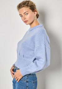 Jasnoniebieski sweter dziany z okrągłym dekoltem, prążkowanym wykończeniem i balonowymi rękawami. Noszony z dżinsami o wysokim stanie. Teksturowany detal z włóczki.