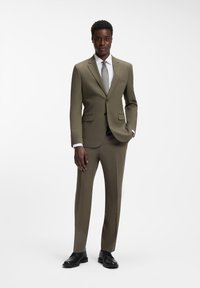 Mann posiert in olivgrünem Anzug mit weißem Hemd, grauer Krawatte und schwarzen Lederschuhen vor weißem Hintergrund.