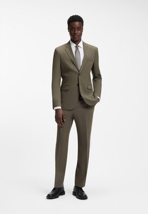 Mann posiert in olivgrünem Anzug mit weißem Hemd, grauer Krawatte und schwarzen Lederschuhen vor weißem Hintergrund.